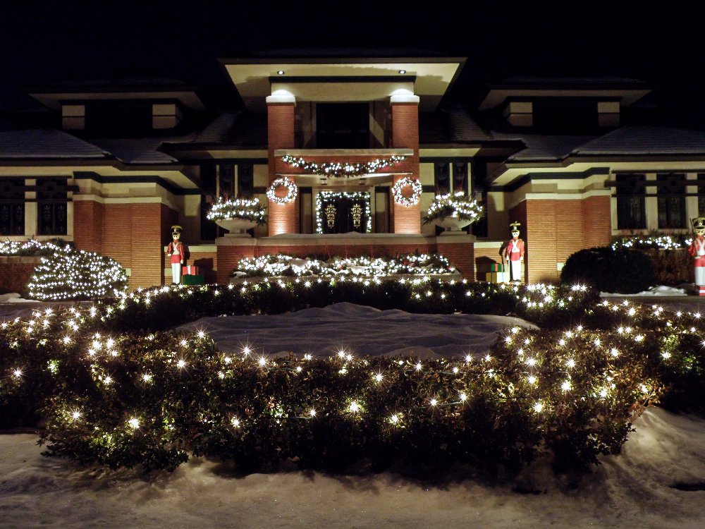 snow covered front yard with lights strung up on bushes and facade