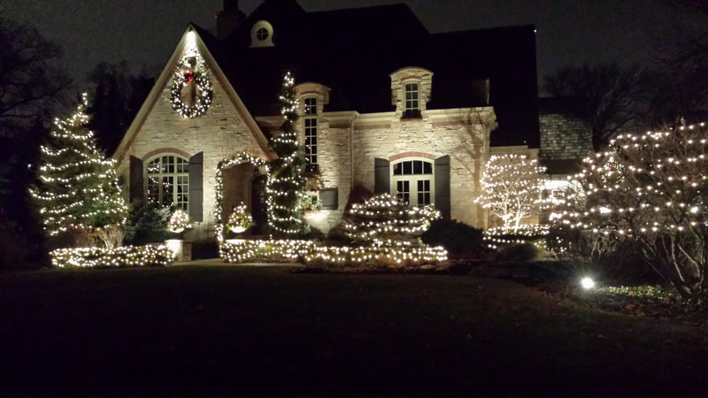 front of large home with lights strung up on all shrubs and bushes